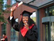 School graduate woman in academic gown and hat looking at the camera with happy smile. Happy multiracial girl rejoicing near her university or school. Graduation concept