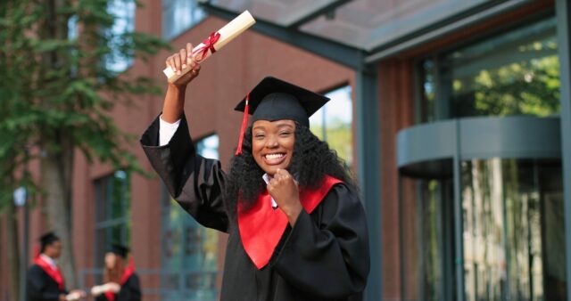 School graduate woman in academic gown and hat looking at the camera with happy smile. Happy multiracial girl rejoicing near her university or school. Graduation concept